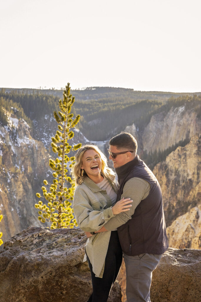 Engaged couple laughing in front of the iconic Lower Falls in Yellowstone at sunset.