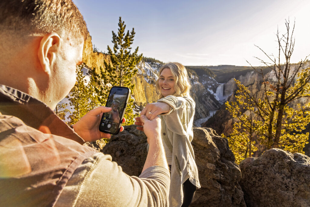 Ashlyn showing off her engagement ring while Kasey photographs it with a cell phone in Yellowstone. Scenic Yellowstone proposal photo featuring the couple and waterfall at sunset.
