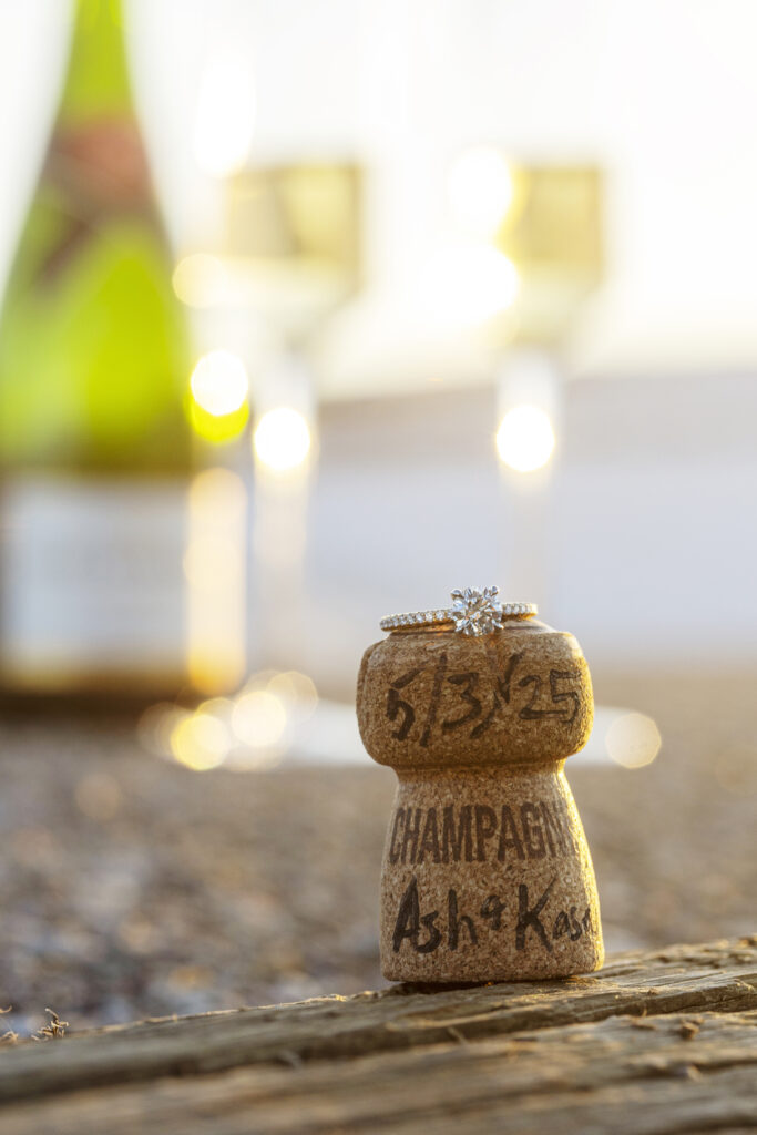 Engagement ring resting on champagne cork engraved with Kasey & Ashlyn’s names and proposal date.