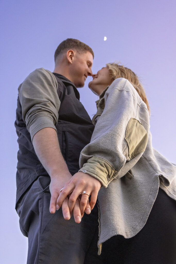Engaged couple laughing and looking at each other with the moon behind them.