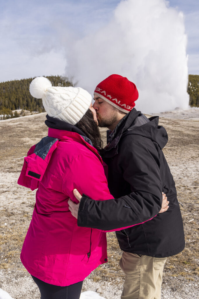 Engaged couple sharing a kiss with Old Faithful erupting in the background during their winter proposal in Yellowstone.