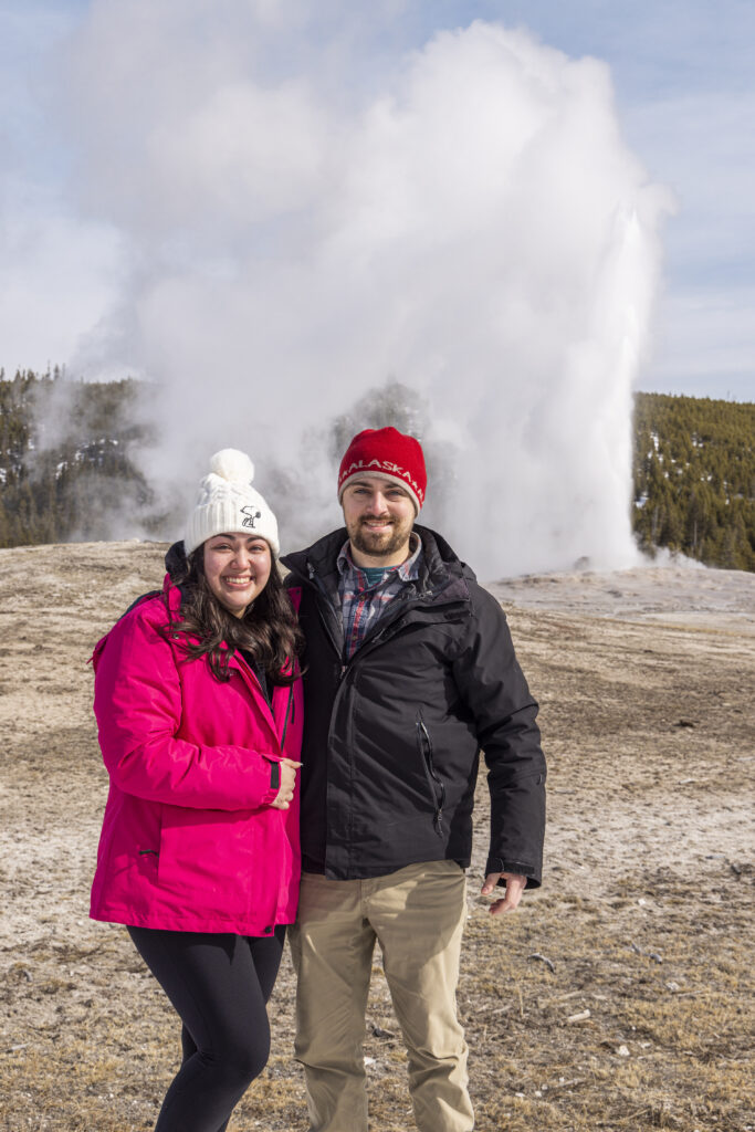 Portrait of the couple standing with Old Faithful erupting behind them.