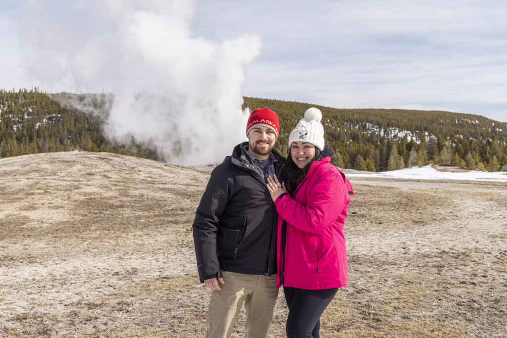 Engaged couple smiling at the camera with Old Faithful erupting in the background during their surprise proposal in Yellowstone.