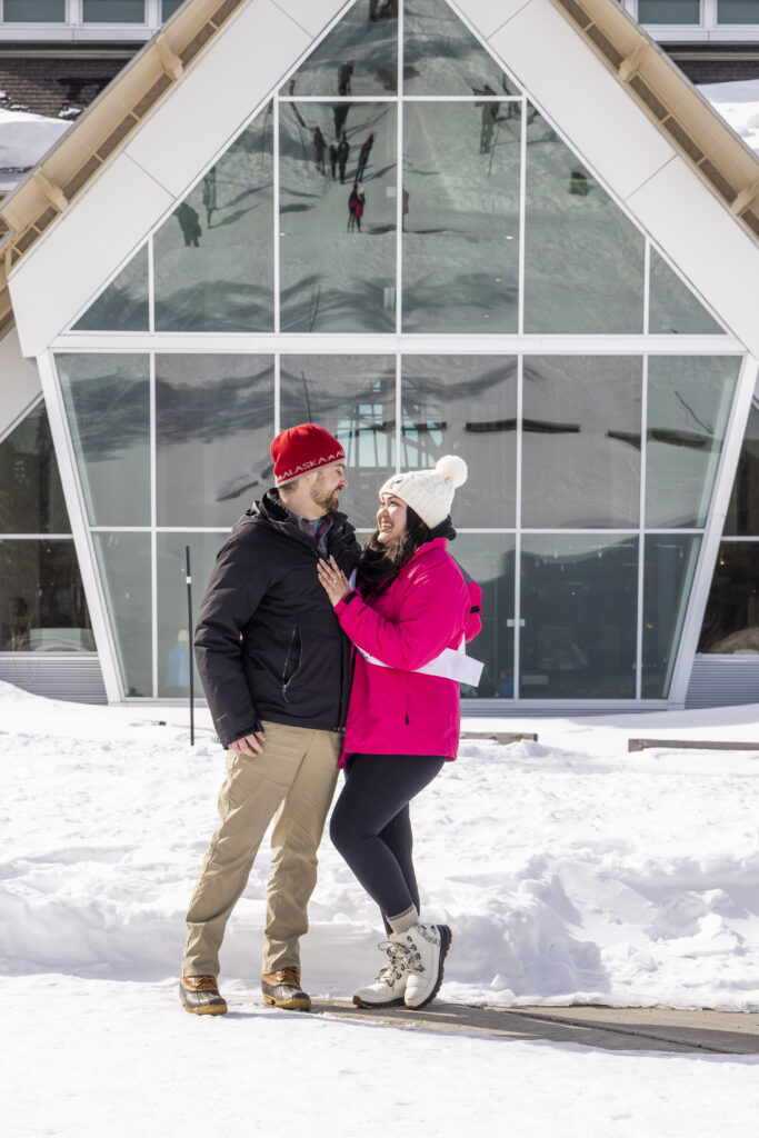 Engaged couple posing in front of the Old Faithful Visitor Education Center with large glass windows reflecting the snowy Yellowstone landscape.