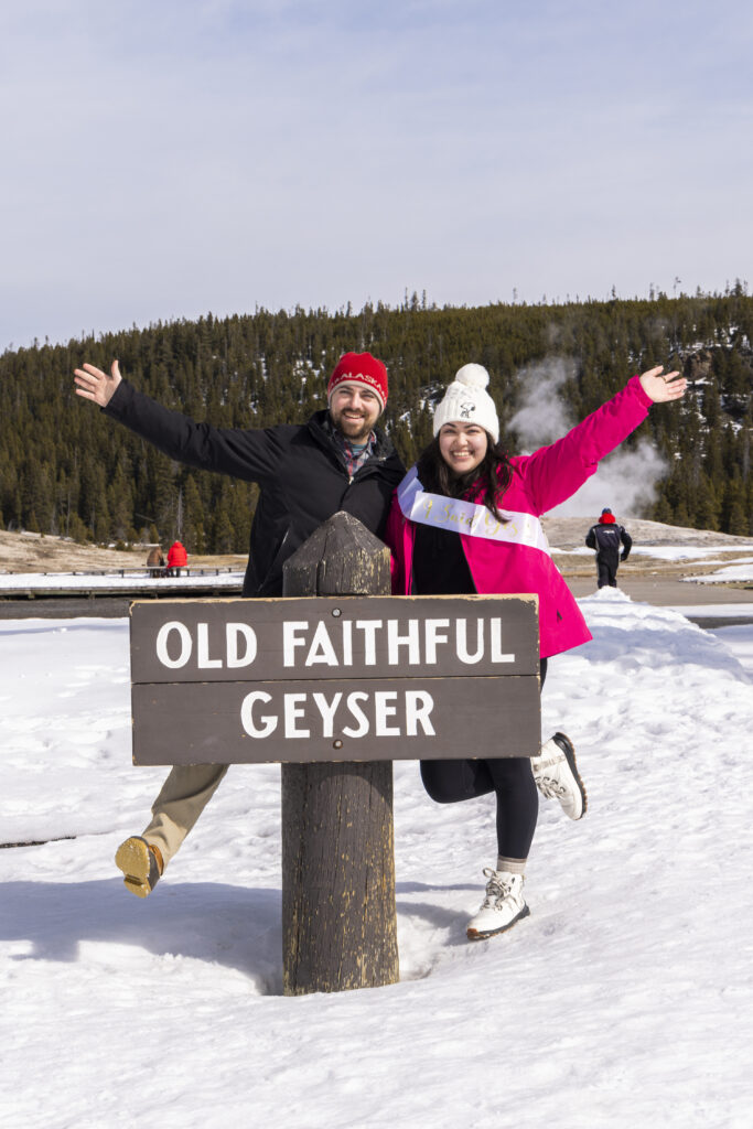 Keith and Jill standing behind the Old Faithful sign, beaming with excitement moments after their surprise proposal in Yellowstone.