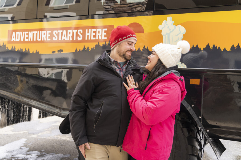 Engaged couple smiling in front of a snowcoach in Yellowstone with the words 'Adventure Starts Here' on the side.